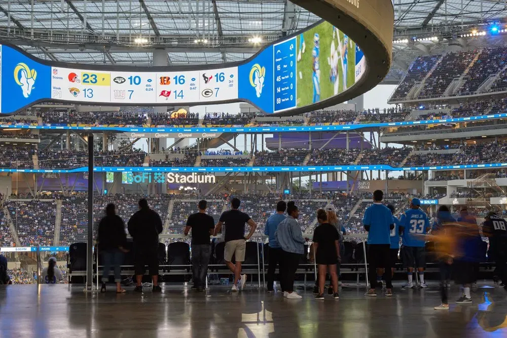 Fans enjoying a football game at SoFi Stadium with a prominent video board and packed seating. Fans inside SoFi Stadium watching a football game, with the large oval video board displaying scores and game highlights. The stadium is filled with spectators across multiple levels.