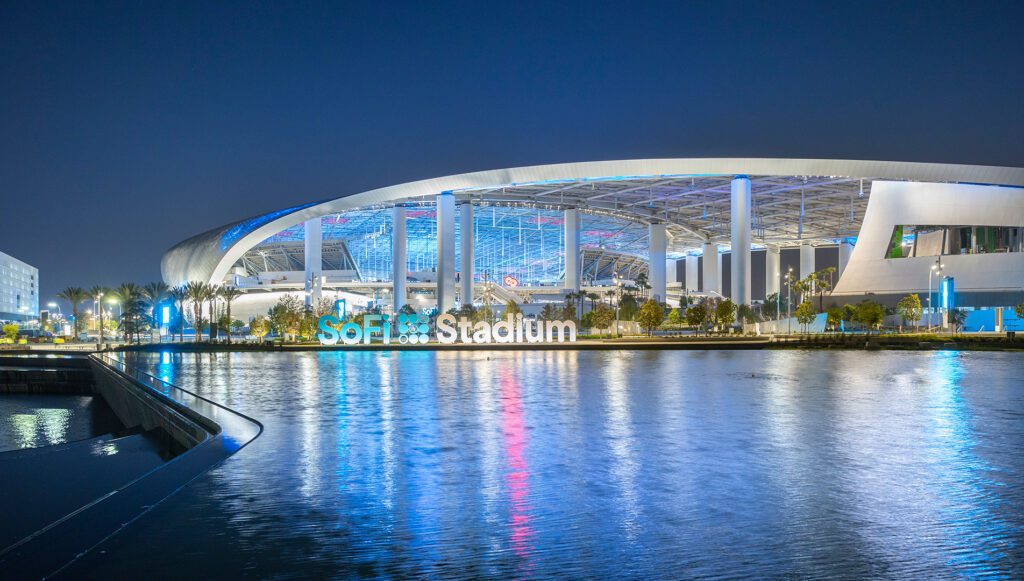 SoFi Stadium at night with reflections on the water. A nighttime view of SoFi Stadium, illuminated with blue and white lights, reflecting on the water in the foreground. The modern architectural design of the stadium is highlighted against the dark sky.