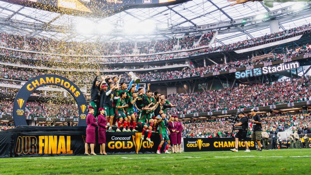 Soccer team celebrates Gold Cup victory at SoFi Stadium with confetti and fans. A triumphant soccer team celebrates winning the Concacaf Gold Cup at SoFi Stadium. The players stand on a stage under a large "Champions" arch while confetti rains down, surrounded by a cheering crowd in the stadium.