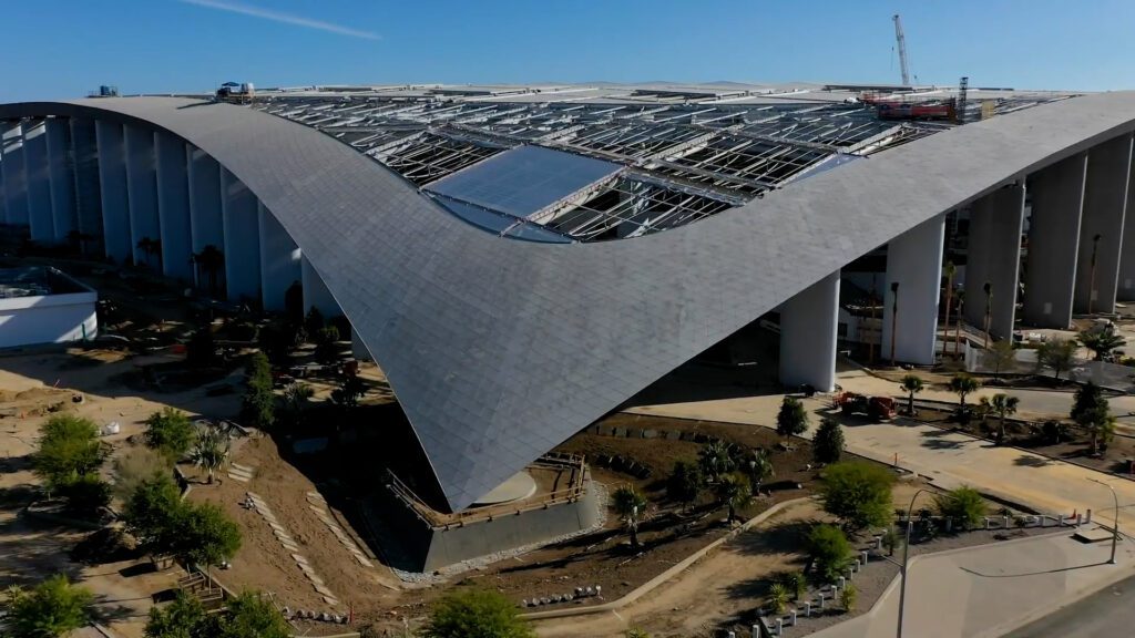 SoFi Stadium under construction with visible curved roof design and landscaping. A close-up aerial view of SoFi Stadium during its construction, showcasing the curved roof structure, open framework, and surrounding landscaping under development.