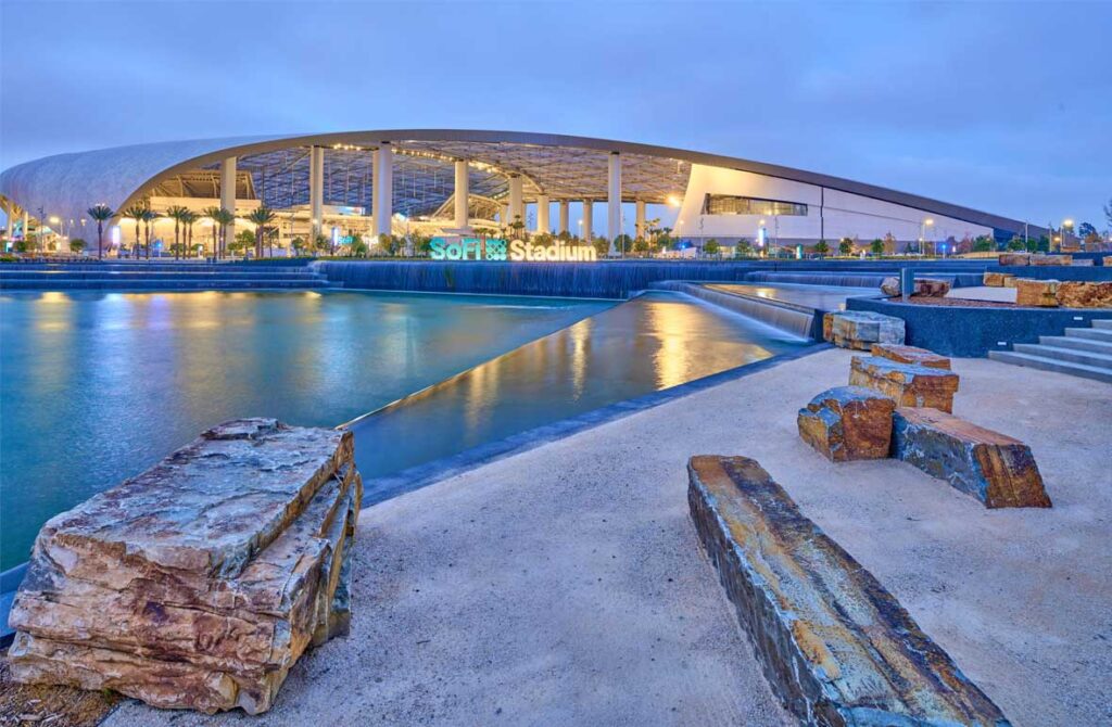 Evening view of SoFi Stadium with reflections on water and landscaping. A scenic evening view of SoFi Stadium, illuminated with lights reflecting off the water in the foreground. The modern architecture is complemented by landscaped surroundings and a stone seating area.