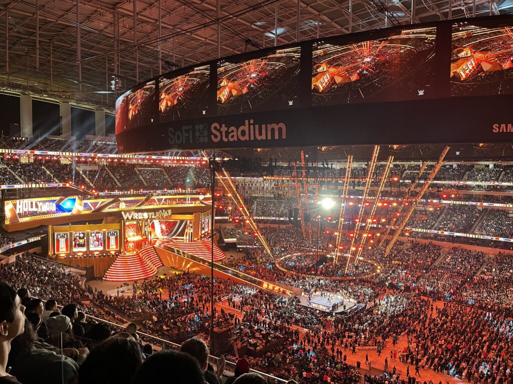Wrestling event at SoFi Stadium with a packed audience and dynamic stage lighting. A vibrant view of a wrestling event at SoFi Stadium, with a packed crowd and colorful stage lighting. The large video board displays the event, and the arena is filled with excitement and energy.