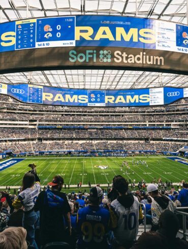 A wide-angle view of a football game at SoFi Stadium, with fans in the foreground wearing Los Angeles Rams jerseys. The massive scoreboard displays "Rams" and game details, while the field and seating areas are filled with spectators.