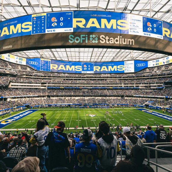 A wide-angle view of a football game at SoFi Stadium, with fans in the foreground wearing Los Angeles Rams jerseys. The massive scoreboard displays "Rams" and game details, while the field and seating areas are filled with spectators.